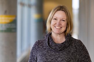 Kelly Muir smiling, wearing a grey turtleneck sweater in an airy modern building